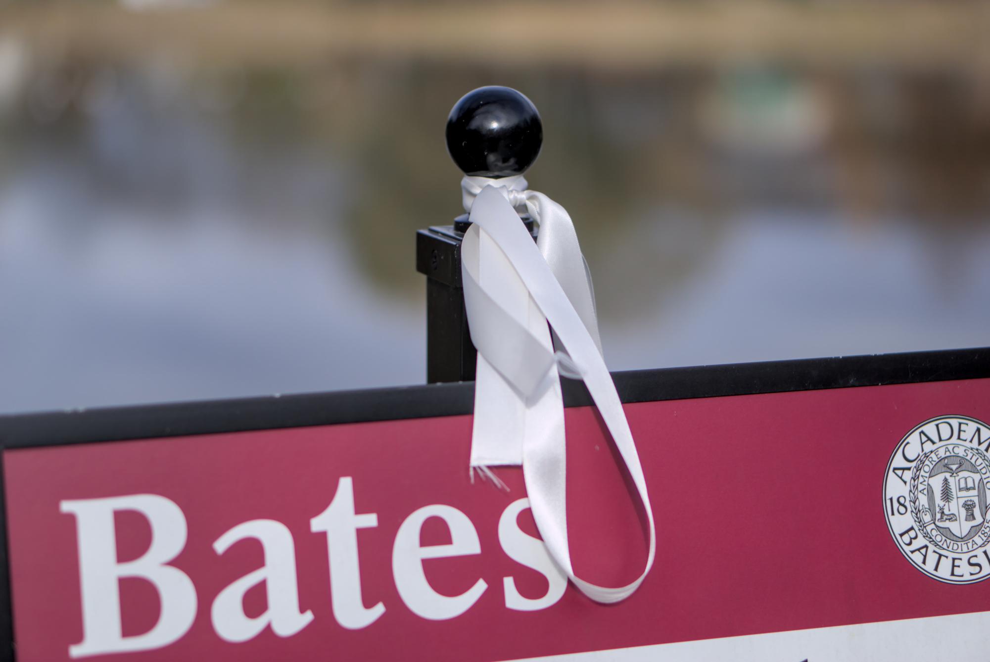 A white ribbon in memory of the victims of the Lewiston mass shooting hangs on a sign on campus. Ribbons were distributed in bundles of 18 and will hang for 18 days to commemorate the 18 people killed on Oct. 25. Noah Skinner/The Bates Student.